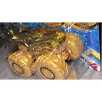 A gold monster truck toy in clear blister packaging, with large textured wheels and a shiny metallic gold body. The package backing shows blue and yellow graphics, and a small red logo is visible in the lower right corner.